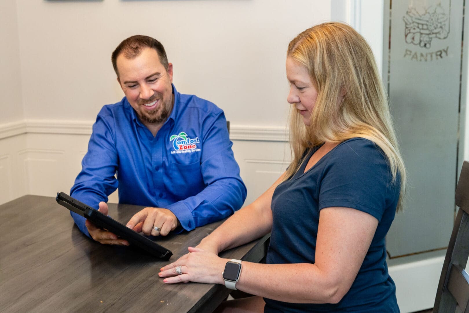 Free AC Estimate A HVAC consultant and homeowner sitting at a table with an iPad reviewing a replacement proposal.
