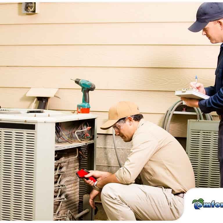 Two HVAC service technicians are analyzing a heating and cooling condenser, inspecting its components to ensure efficient operation