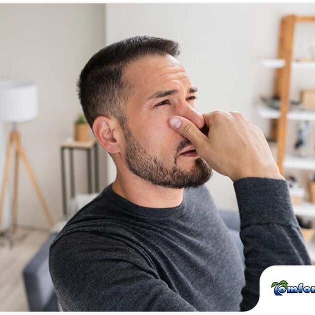 A man is holding his nose while looking at the camera, indicating discomfort due to poor indoor air quality in the home