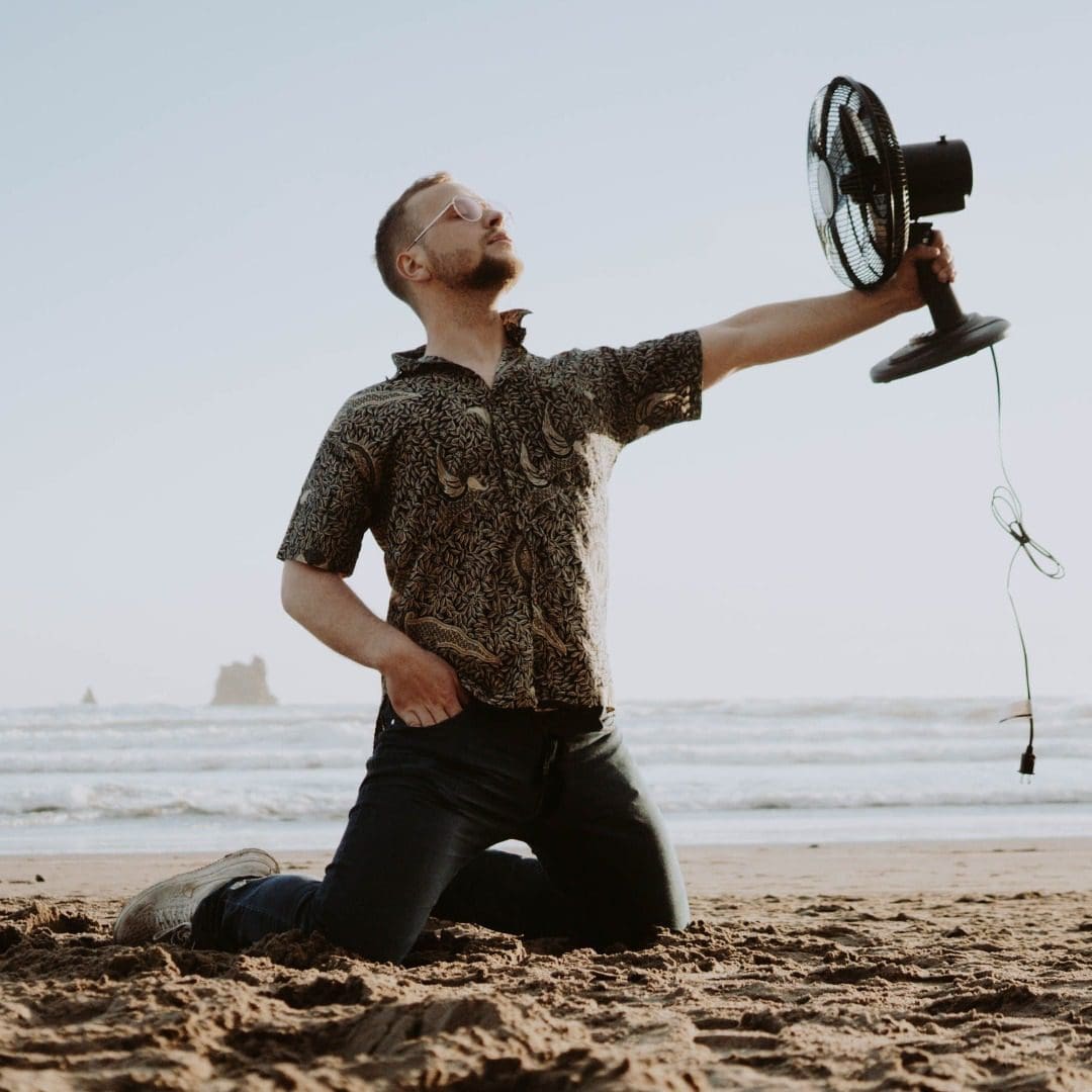 A man is playing with a fan on the beach that is not plugged in, likely pretending to use it in the breeze or as a playful prop